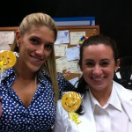 Two women smiling with Sudsies smiley cookies in front of a message board